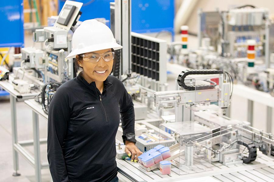 An individual wearing a white hard hat and safety goggles smiles while standing in front of machinery